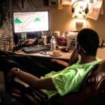 Man relaxing in a cozy home office with a computer setup, lamp, and chess set, enjoying some leisure time.